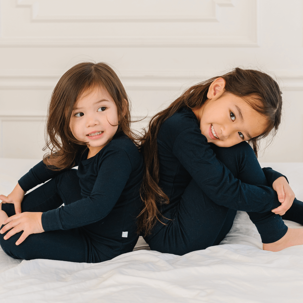 Two young girls in matching navy blue outfits sitting on a white bed.