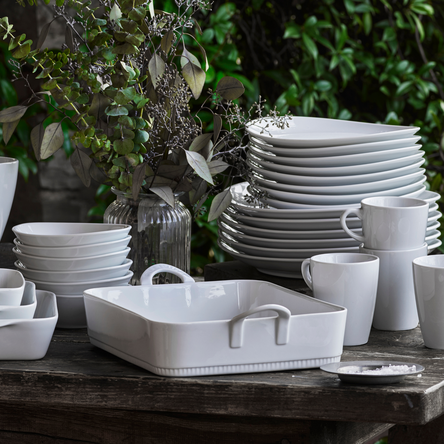 White ceramic tableware including bowls, plates, and mugs on a wooden table with greenery in the background.