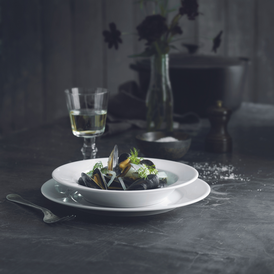 Mussels in a white bowl on a dark surface with a glass of white wine and decorative elements.