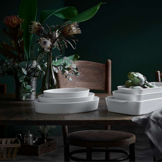 Many White ceramic dishes on a wooden table with a dark background and decorative plants.