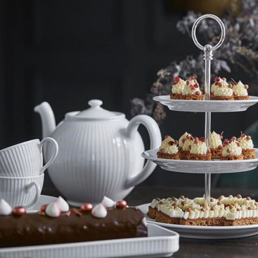 Tea set with a teapot, cups, and a three-tiered stand with small desserts on a dark background.