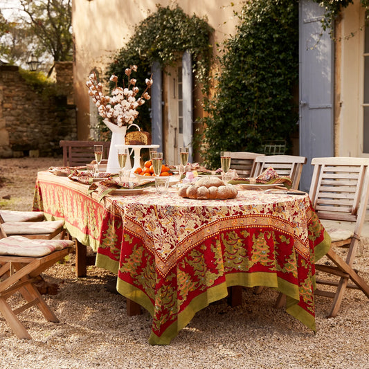 A tablecloth with a red and green Christmas-themed design is laid out on a table outdoors, with a decorated table in the background and a building in the background.