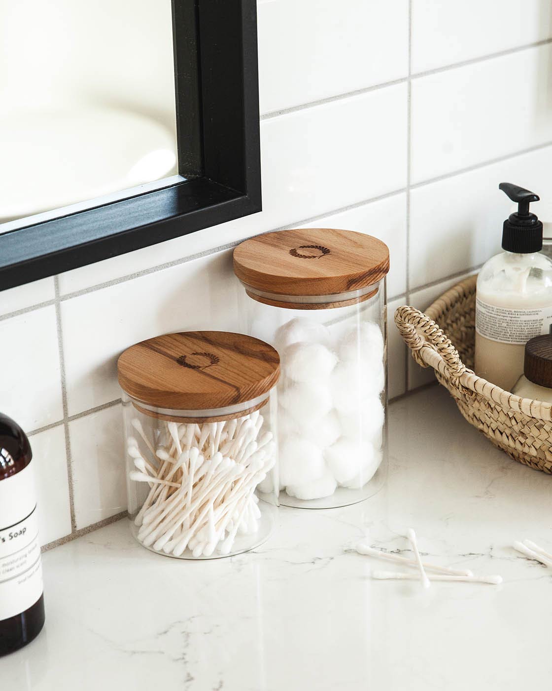 Bathroom counter with glass jars with wooden lids containing cotton swabs and cotton balls, next to a bottle of soap.