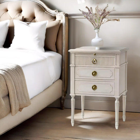 White nightstand with gold knobs next to a bed with white bedding and beige pillows.