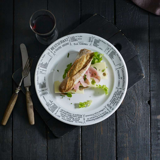 Plated dish with a sandwich on a decorative plate, accompanied by cutlery and a glass of red wine on a dark wooden table.