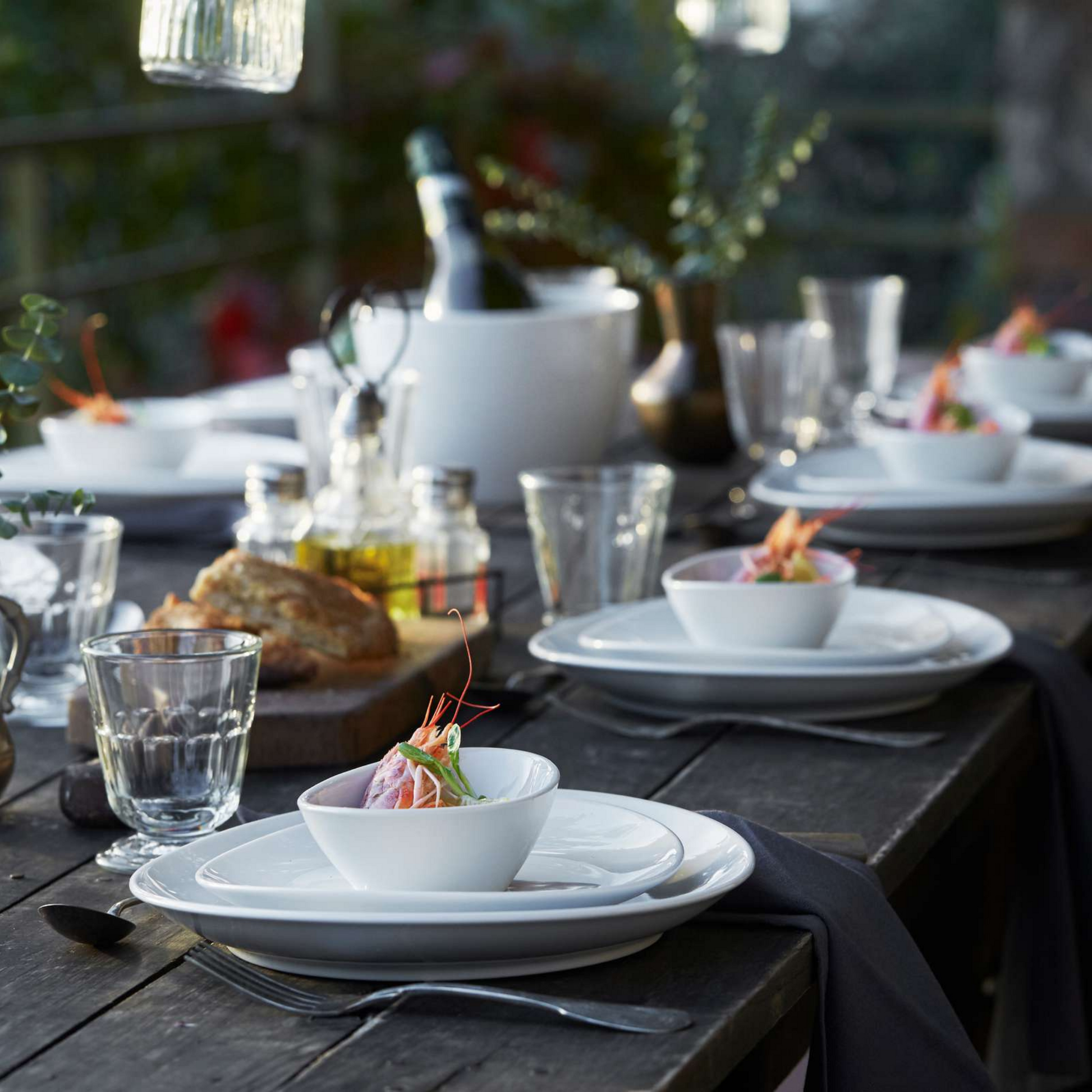 Outdoor dining setup with plates, bowls, and glasses on a wooden table.