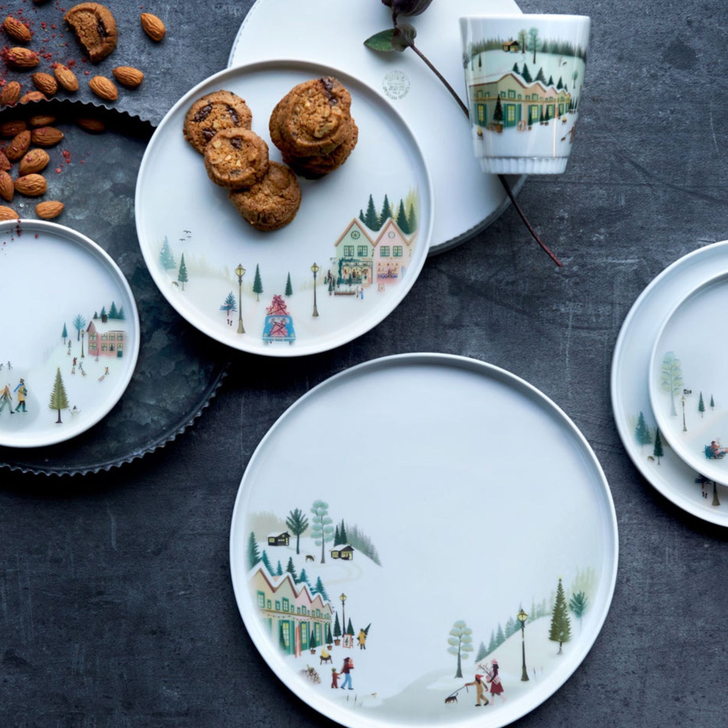 Decorative plates with winter scene design on a dark surface with cookies and nuts.