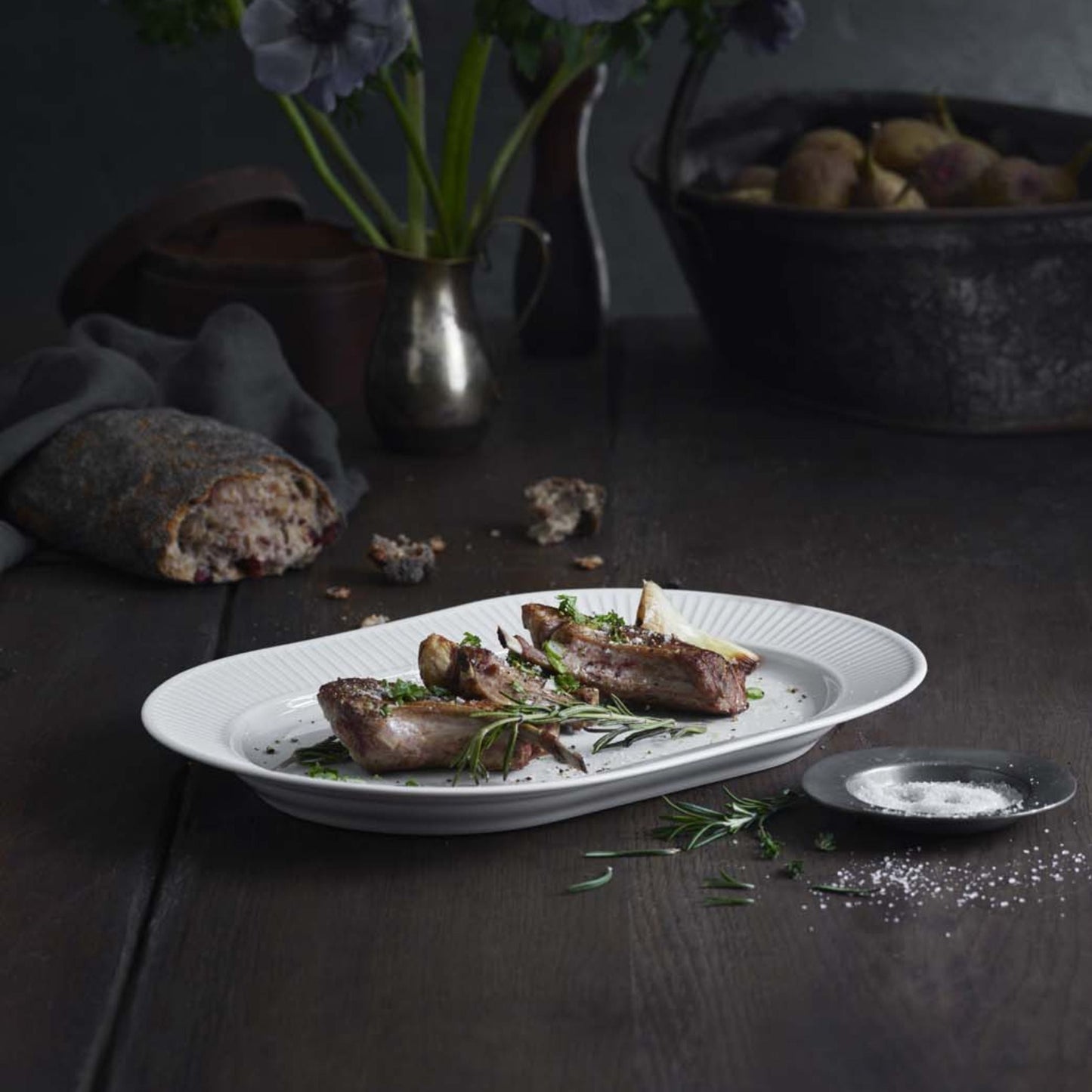 Plated dish of roasted meat with herbs on a dark wooden table, accompanied by bread and a vase of flowers.