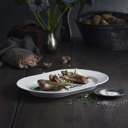 Plated dish of roasted meat with herbs on a dark wooden table, accompanied by bread and a vase of flowers.