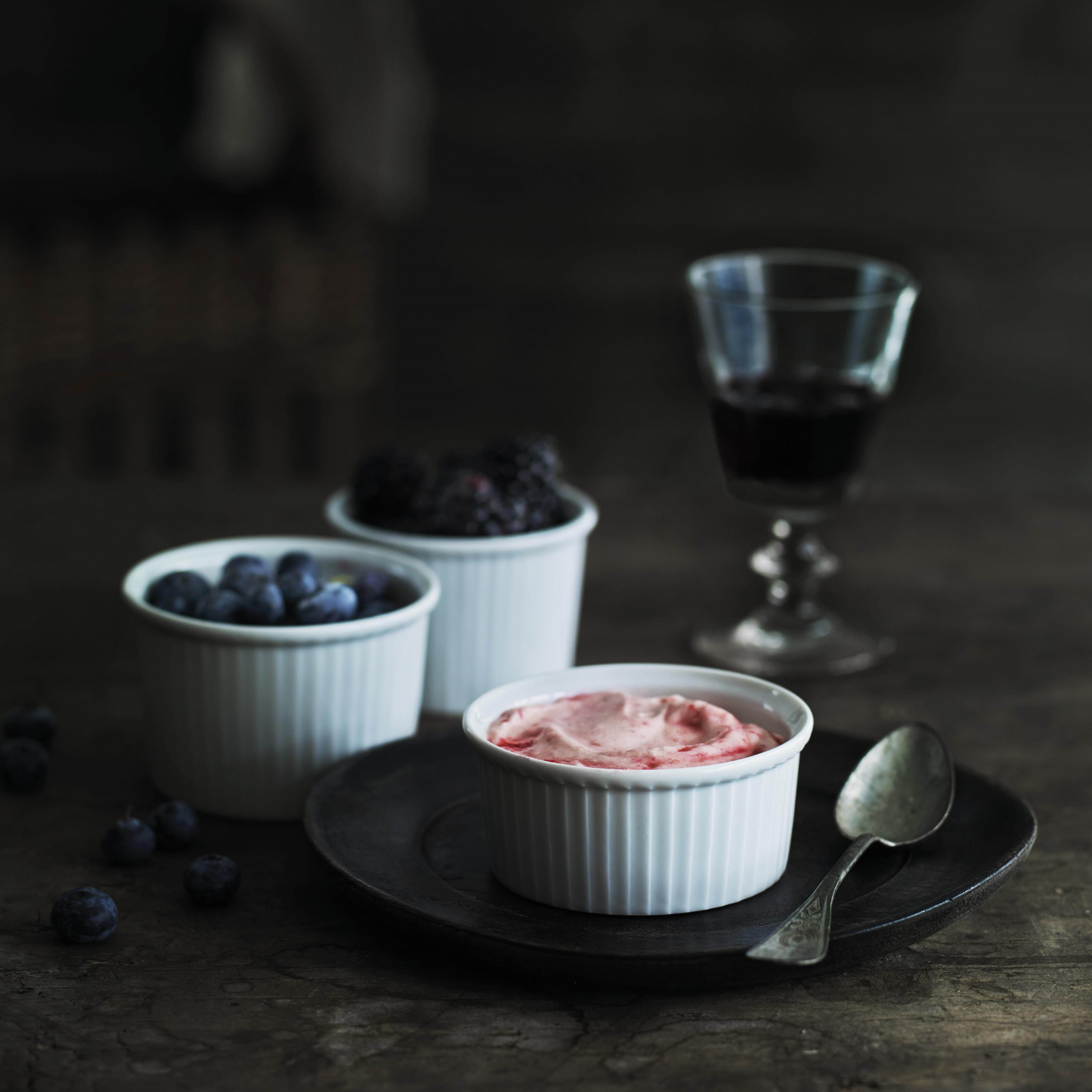 Three white ramekins with different desserts on a dark surface, accompanied by a glass of dark liquid.