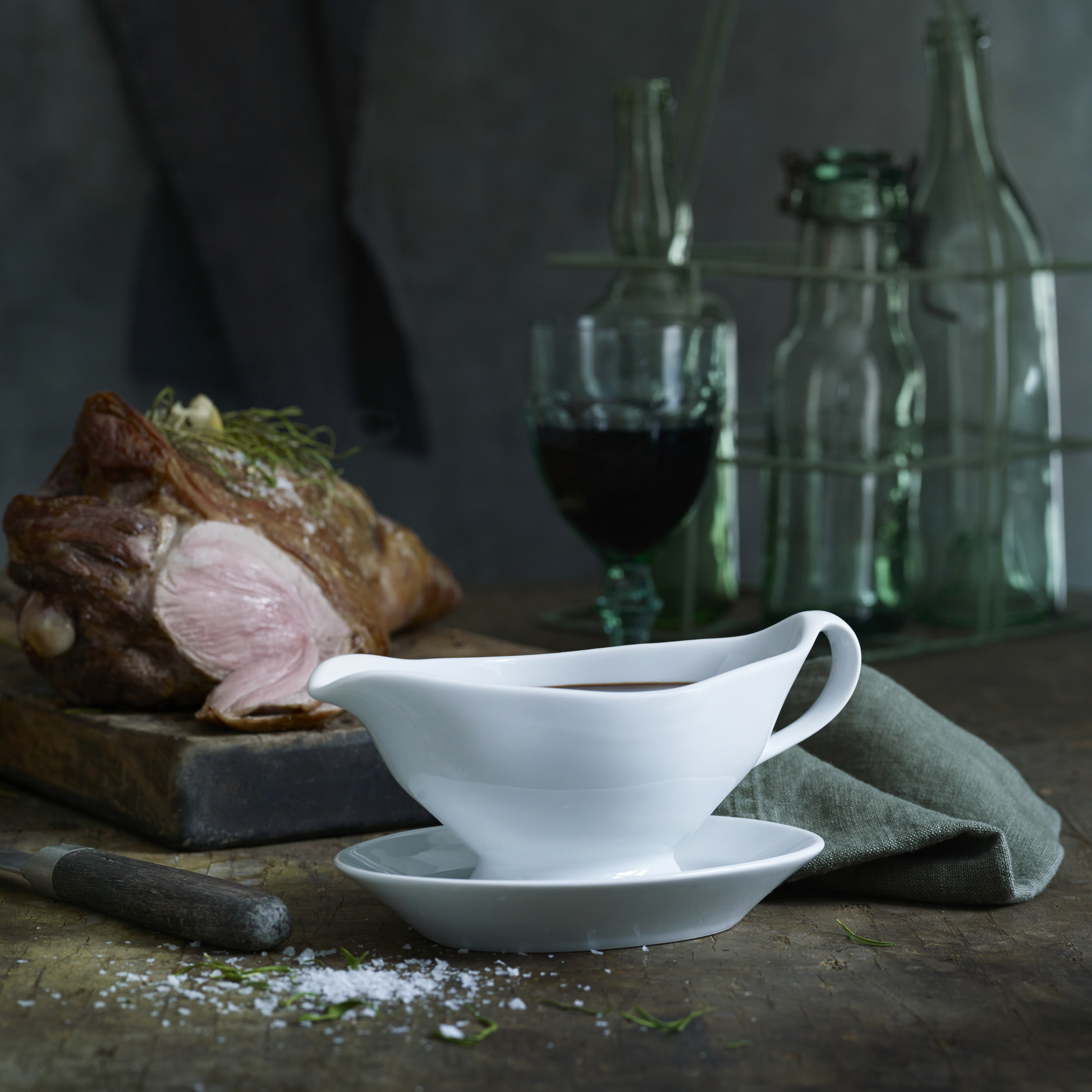 White gravy boat and bowl on a rustic wooden table with a roasted meat dish and bottles in the background.