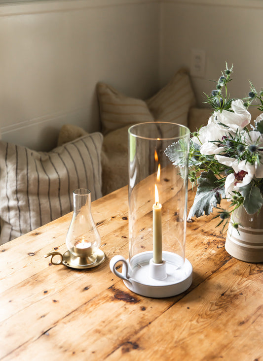 Candle in a glass holder on a wooden table with flowers and pillows in the background