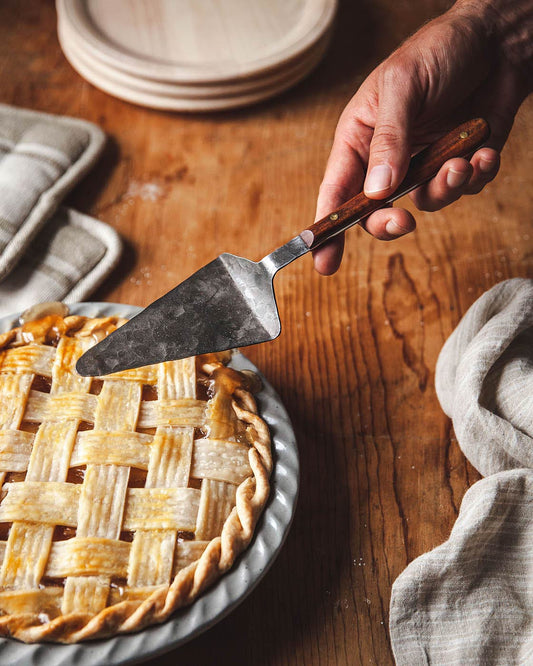 Hand using a pie server to cut into a lattice-topped pie on a wooden table.