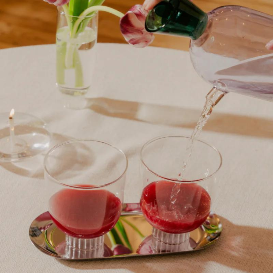 Person pouring liquid into two glasses on a tray with a vase in the background