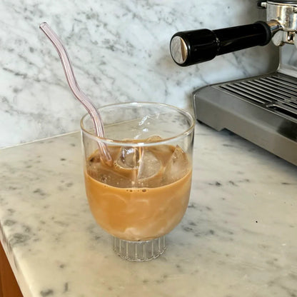 Glass of iced coffee with a straw on a marble countertop next to an espresso machine.