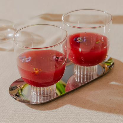 Two red cocktails with floral garnishes in clear glasses on a mirrored tray.