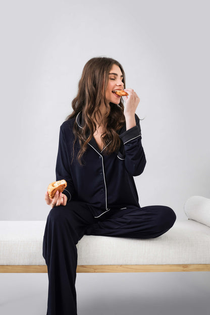 Woman in black pajamas eating a donut on a white bench with a light gray background