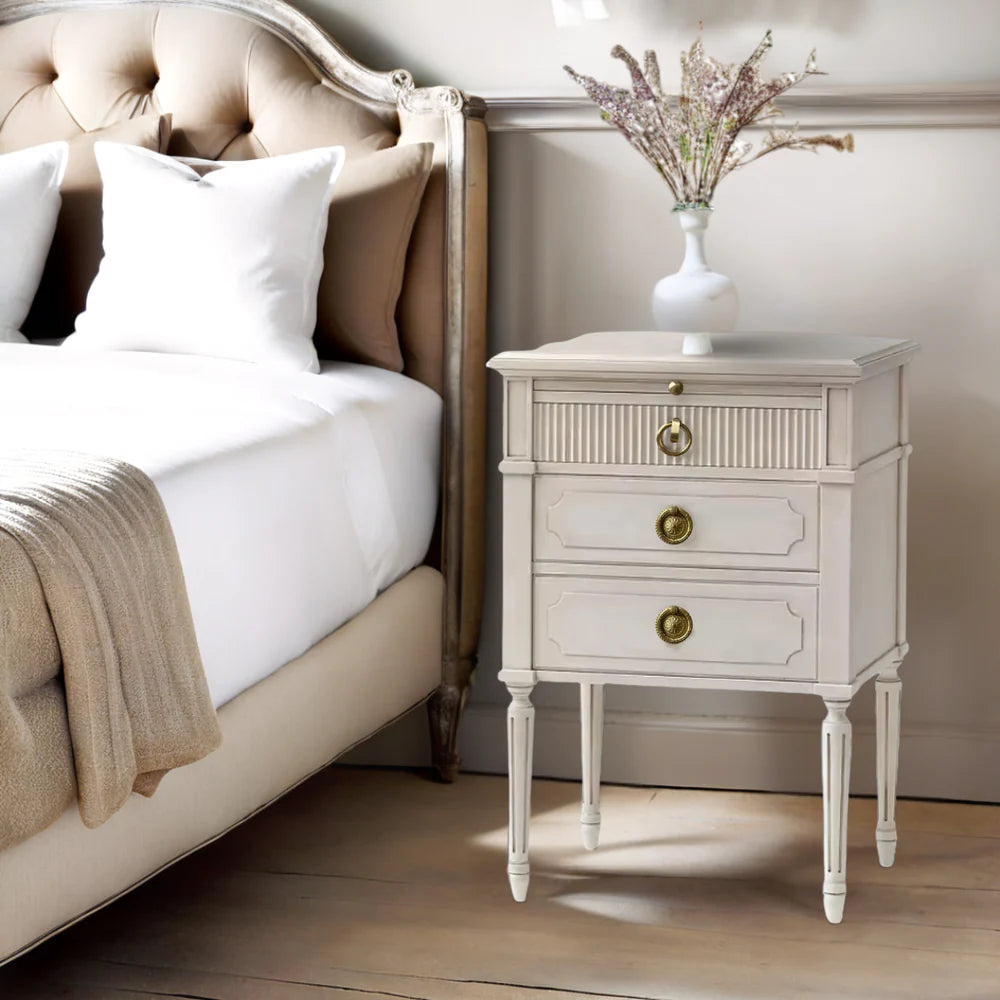 White nightstand with gold knobs next to a bed with white bedding and beige pillows.