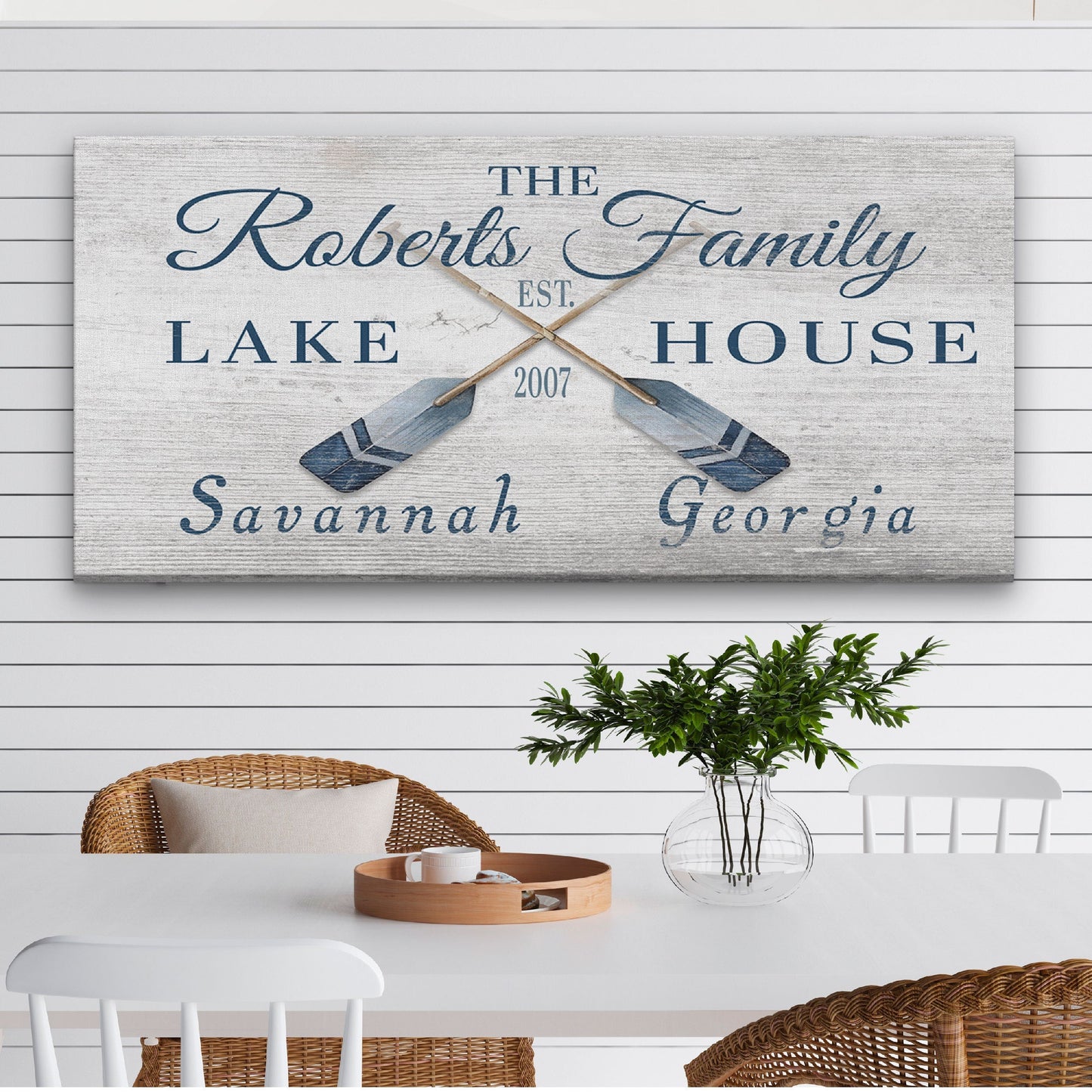 Decorative sign with customizable printing'The Roberts Family Lake House' text and oar design on a white wall above a dining table.