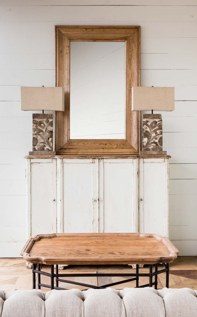 Wooden mirror above a white cabinet with lamps on either side, in a room with a wooden table and gray sofa.