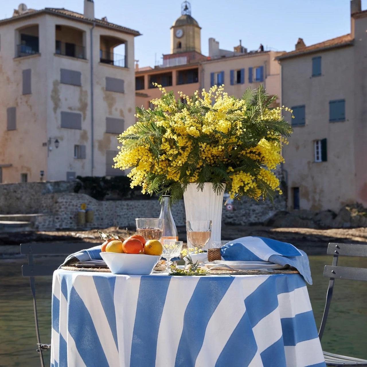 Table setting with yellow flowers and a striped tablecloth by a waterfront with buildings in the background.