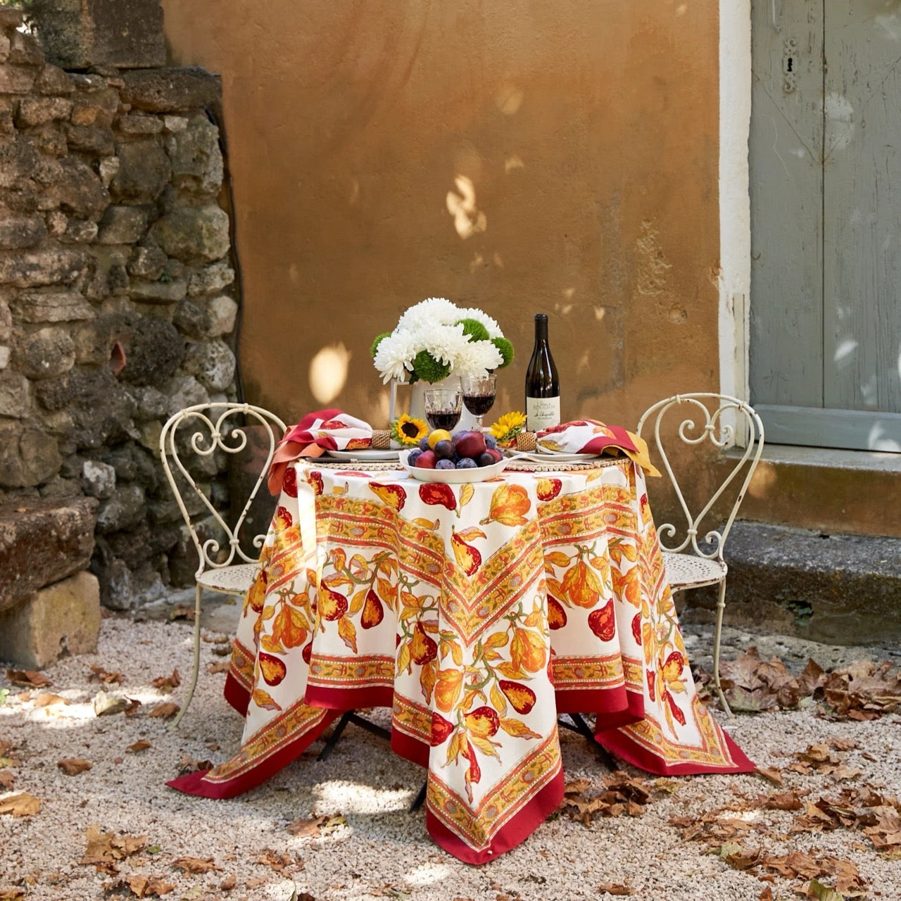 Outdoor table setting with a decorative tablecloth, wine glasses, and flowers against a stone wall.