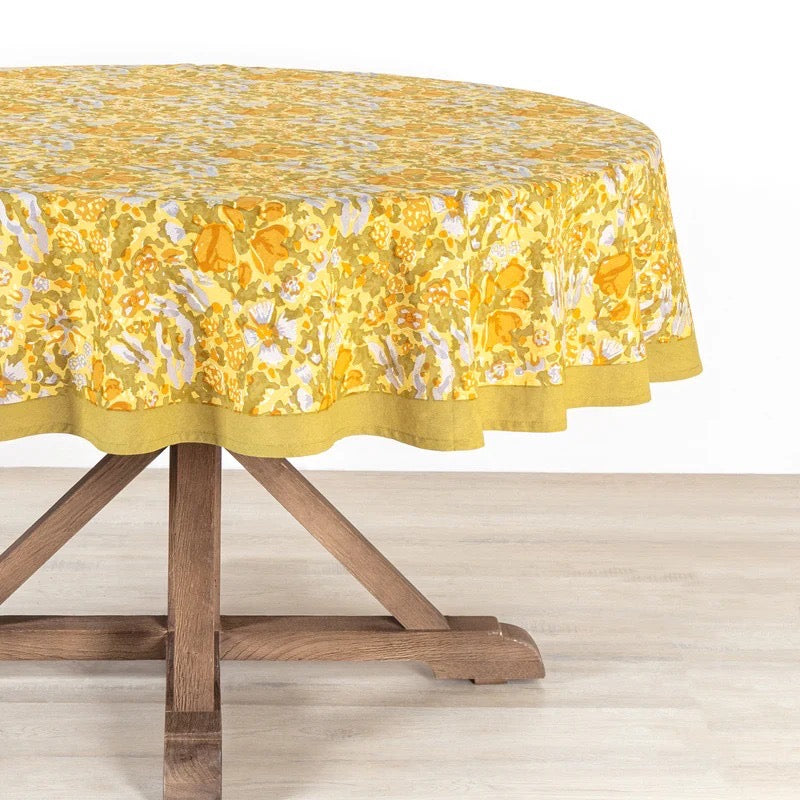 Round table with a yellow floral tablecloth on a wooden table.