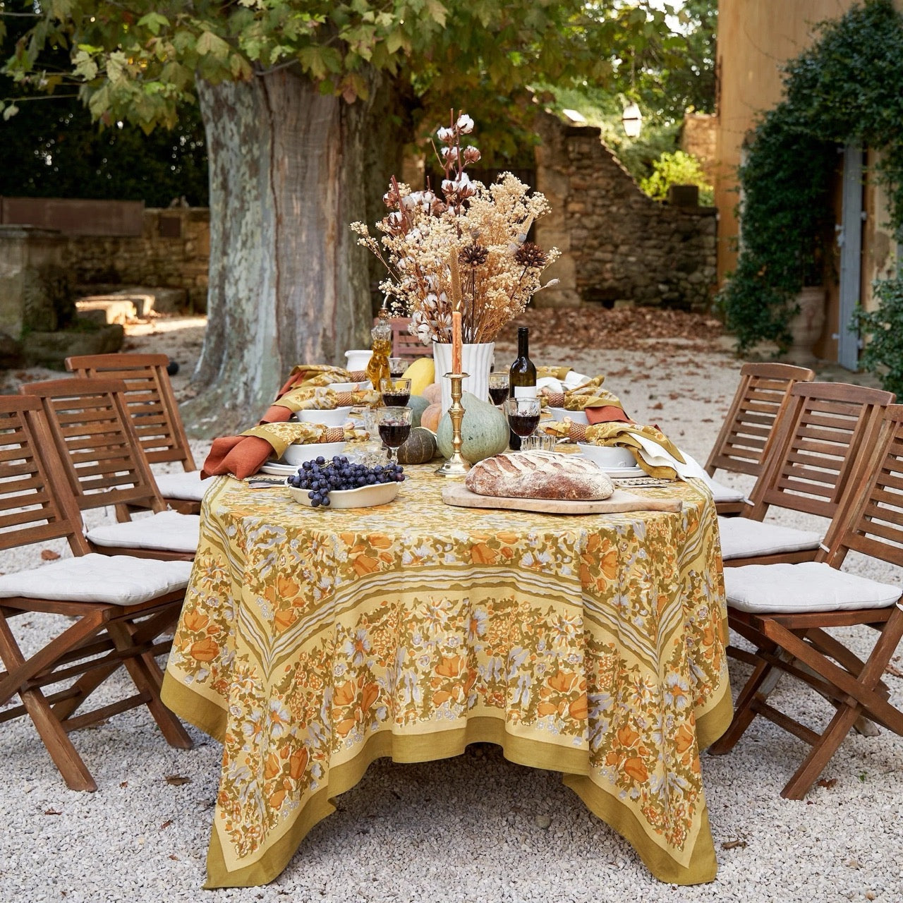 Outdoor dining table set with a floral tablecloth, wine, bread, and fruit in a rustic setting.